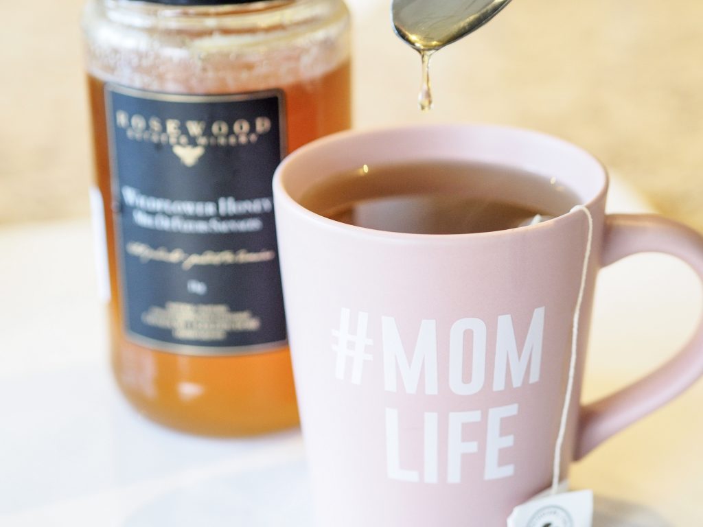 Traditional Medicinals Herbal Teas organic raspberry leaf tea steeping inside pink #momlife teacup with spoon dripping honey into the mug, and a jar filled with organic honey.