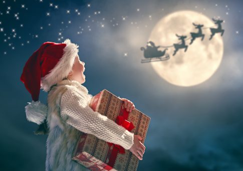 Merry Christmas! Cute little child with xmas present. Santa Claus flying in his sleigh against moon sky. Happy kid enjoy the holiday. Portrait of girl with gifts on dark background.