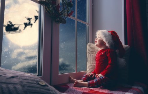 Merry Christmas and happy holidays! Cute little child baby girl sitting by window and looking at Santa Claus flying in his sleigh against moon sky. Room decorated on Christmas. Kid enjoy the holiday.